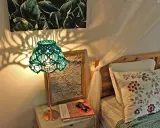 Oświetlona lampa z zielonym, plecionym abażurem, pokazująca, jak zrobić abażur ze sznurka. Cienie rzucane na ścianę tworzą ciekawy wzór.