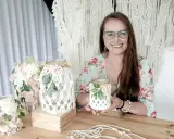 Uśmiechnięta kobieta prezentuje, jak zrobić lampion makrama. W tle widać gotowe ozdoby i materiały.