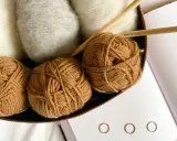 Kłębek brązowej włóczki, białe i szare włókna, druty i książka o hygge. Różne grubości włóczek zachęcają do tworzenia.