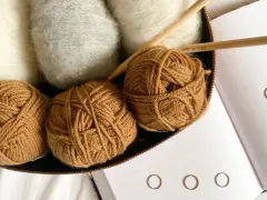 Kłębek brązowej włóczki, białe i szare włókna, druty i książka o hygge. Różne grubości włóczek zachęcają do tworzenia.