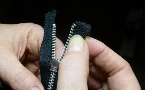 Dłonie próbują naprawić zepsuty zamek w pościeli. Zamek jest czarny, metalowy, z widocznym suwakiem YKK.