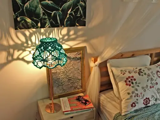 Oświetlona lampa z zielonym, plecionym abażurem, pokazująca, jak zrobić abażur ze sznurka. Cienie rzucane na ścianę tworzą ciekawy wzór.