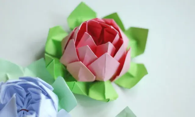 Różowy i niebieski kwiat origami prosty, otoczony zielonymi listkami.