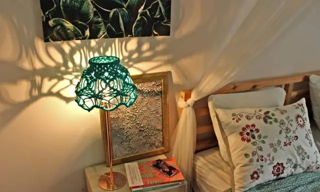 Oświetlona lampa z zielonym, plecionym abażurem, pokazująca, jak zrobić abażur ze sznurka. Cienie rzucane na ścianę tworzą ciekawy wzór.