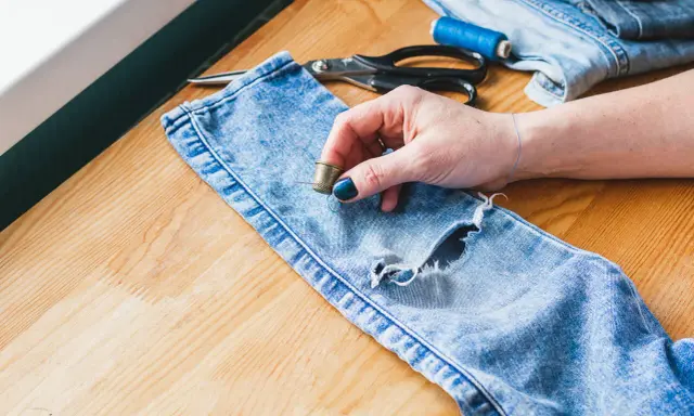 Naprawianie dziur w ubraniach. Dłoń z naparstkiem i igłą przy podartych jeansach, obok nożyczki i nici.
