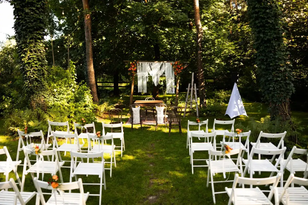Uroczysta ceremonia ślubna w ogrodzie. W centralnym punkcie znajduje się drewniany łuk ozdobiony piękną makramą ślubną, a przed nim st&oacute;ł dla nowożeńc&oacute;w. Wok&oacute;ł ustawiono rzędy białych krzeseł dla gości.