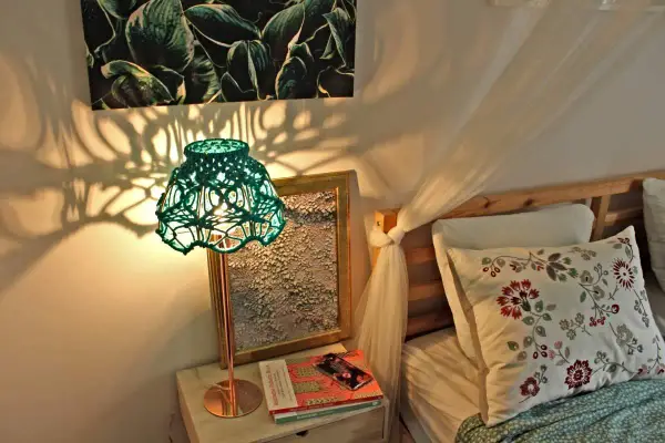Oświetlona lampa z zielonym, plecionym abażurem, pokazująca, jak zrobić abażur ze sznurka. Cienie rzucane na ścianę tworzą ciekawy wzór.